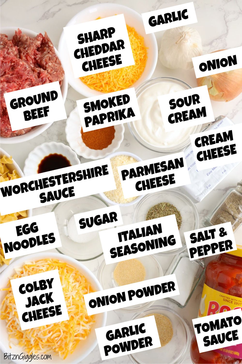 Ground beef, egg noodles, cheeses, sour cream, cream cheese, tomato sauce, onion, garlic and seasonings arranged on a countertop.